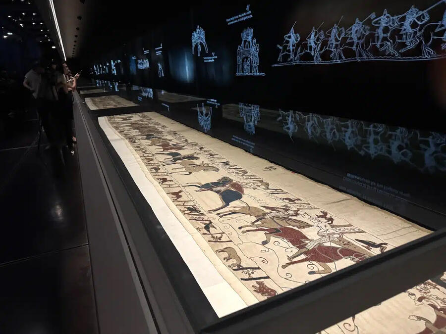 The Bayeux Tapestry displayed horizontally beneath the glazed hoods of the 70-metre showcase.